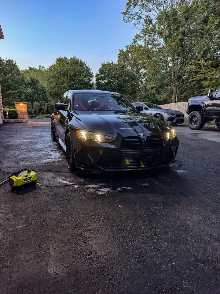 A black BMW M3 being washed in a driveway, pressure washer in the foreground